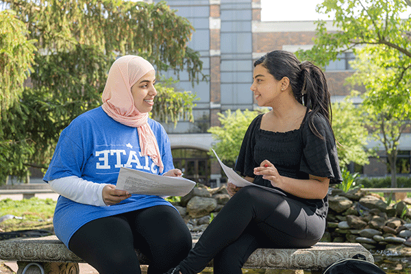 Two females sit on a bench talking while holding papers